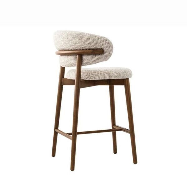 The Caelina bar stool, seen from the right side, displaying the ergonomic curvature of the backrest and the thick, cushioned seat. The wooden frame, finished in a light brown tone, features clean lines and a minimalist design, with the footrest bar visible between the front and back legs.