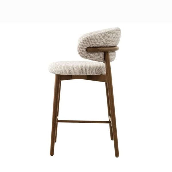 he Caelina bar stool, viewed from the left side, emphasizing the gentle curve of the backrest that wraps around for enhanced support. The wooden legs, finished in light brown, are connected by a footrest bar, adding both stability and functionality. The light, textured fabric of the upholstery adds a touch of elegance to the overall design.