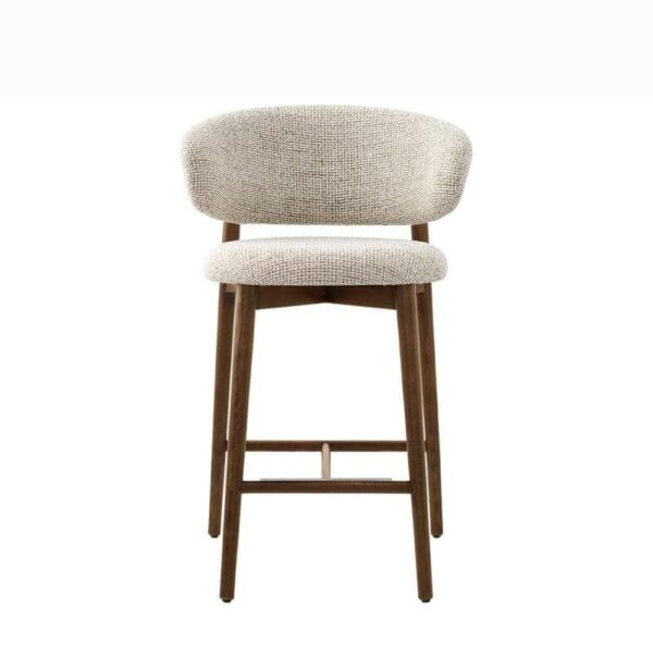 The Caelina bar stool, captured from a direct front view, highlighting its sophisticated design. The rounded backrest and plush seat, both upholstered in a light, textured fabric, sit atop slender wooden legs that taper slightly towards the floor. A footrest bar connects the front legs for added support and comfort.