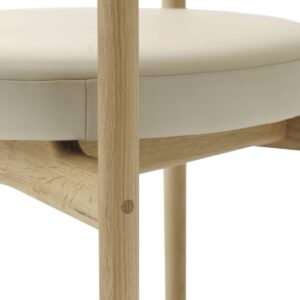 The image shows a detailed close-up of a wooden chair, focusing on the seat area. The seat is upholstered in a smooth, cream-colored fabric, tightly wrapped around a round cushion. The frame of the chair is made from light-toned wood, exhibiting a natural grain and texture. A prominent dowel joint is visible on the wooden leg, highlighting the craftsmanship and sturdy construction of the chair. The minimalist and clean design suggests a modern aesthetic, emphasizing the natural beauty of the materials used.