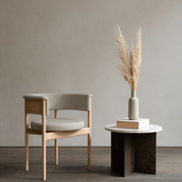 This image features a modern armchair positioned in a minimalist setting. The chair has a sturdy beech wooden frame with a natural finish that accentuates its clean lines and curved armrests. The seat and backrest are upholstered in a subtle grey fabric, offering a soft contrast to the wooden elements. To the right of the chair, a small round table with a marble top and dark wooden base holds a matte grey vase with tall, wispy pampas grass, adding a touch of organic texture to the scene. The background is a simple, light grey wall, enhancing the chair's contemporary aesthetic.