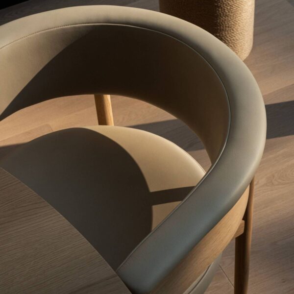 a top-down view of a modern wooden chair, showcasing its elegant design. The chair features a curved backrest upholstered in smooth, beige fabric, matching the cushioned seat. The wooden frame is light-toned, and the craftsmanship is evident in the seamless joinery and smooth finish. The lighting creates soft shadows, emphasizing the gentle curves and contours of the chair. The background includes a wooden floor, harmonizing with the natural aesthetic of the chair. This setting highlights the chair's sophisticated and contemporary style.