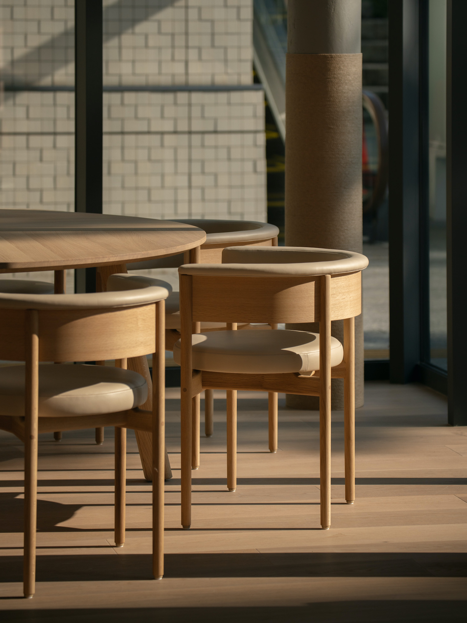 The image shows a selection of Circa Dining Armchairs in a well-lit interior space. The chairs feature curved wooden frames and padded seats in a neutral color, tucked under matching round wooden tables. The flooring is wooden, and the natural light highlights the smooth finish of the wood, suggesting a chic café or contemporary dining area.