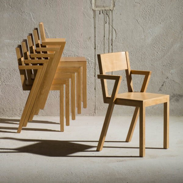 he Cassia Stacking Light Wood Dining Armchair by Kersetna is shown in a minimalist setting. Several chairs are stacked together on the left, showcasing their stackable design, while one chair stands separately on the right. The chairs have a contemporary style with angular armrests and a light wood finish. The background is a textured concrete wall, providing a contrasting backdrop to the smooth, sleek appearance of the chairs.