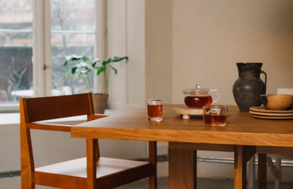 cozy dining scene featuring the Granny Oak Dining Table by Kersetna, set in a bright room with large windows overlooking a winter scene. The table is adorned with a tea set, including a glass teapot and two glasses of tea, alongside a large antique ceramic vase and a stack of plates, creating an inviting and homely atmosphere. The natural wood finish of the table and the simplicity of the design emphasize the beauty of the oak wood.