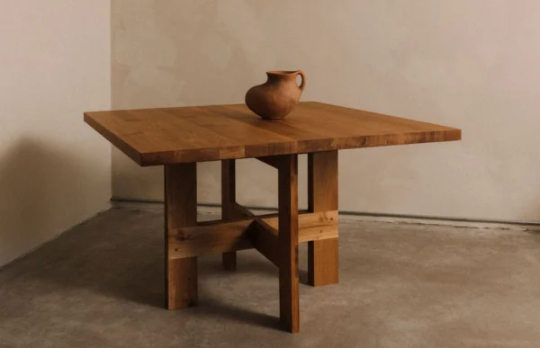 Simplistic yet elegant setting showcasing the Granny Oak Dining Table by Kersetna, displayed in a light-filled room with neutral walls. The table, crafted from oak wood and finished in oil, features a robust square design with a unique interlocking leg structure. A single rustic clay pot rests on the tabletop, accentuating the table’s natural and refined aesthetic