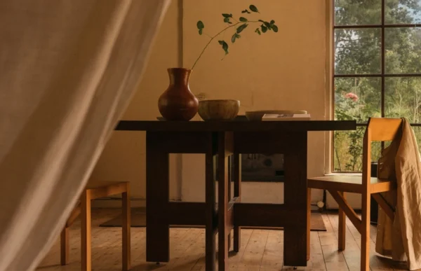 Rustic and warmly lit dining area featuring the Granny Oak Dining Table by Kersetna, crafted from oak wood and finished in oil for a natural look. The sturdy, dark wood table with geometric legs is adorned with a large clay vase and a wooden bowl, enhancing the cozy ambiance. A chair draped with a linen throw and large windows offering a view of greenery outside complement the earthy tones of the setting