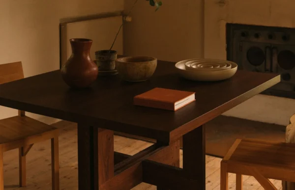 Close-up view of the Granny Oak Dining Table by Kersetna, showcasing the rich dark oak wood finish and oil treatment. The table is set with a large rustic clay vase, a wooden bowl, and a set of ceramic plates, creating an inviting dining setting. The natural light and minimalist decor emphasize the table's robust design and craftsmanship