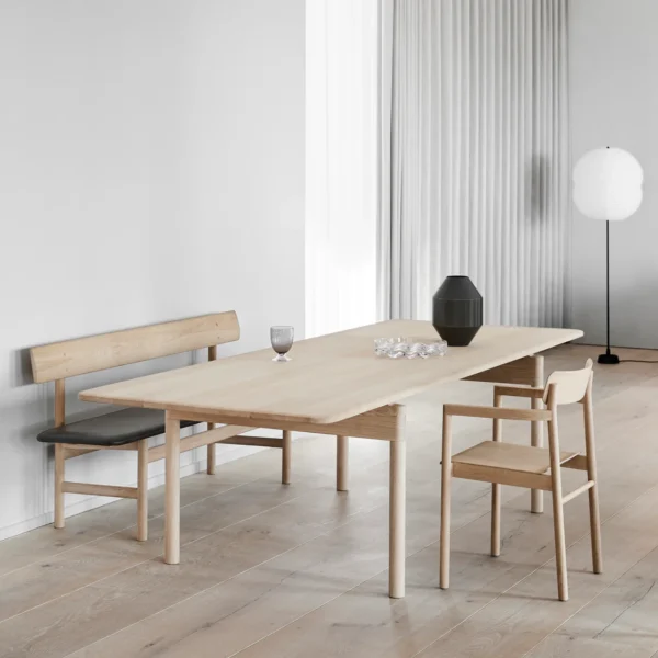 Modern dining area featuring the Visiteur oak wood bench with a black faux leather seat, paired with a minimalist oak dining table and matching chairs. The space is styled with a neutral color palette, light wooden flooring, sheer white curtains, and minimalist decor elements like a sculptural vase and a floor lamp, reflecting Scandinavian design principles of simplicity and natural materials.