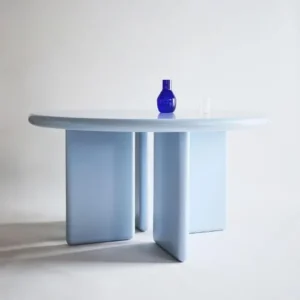 Blau round dining table, by kersetna lebanon, made out of Mdf table top and legs, painted in light blue, on the top of the table a decorative blue glass vase, the setup of the table is in a serene white studio with natural light