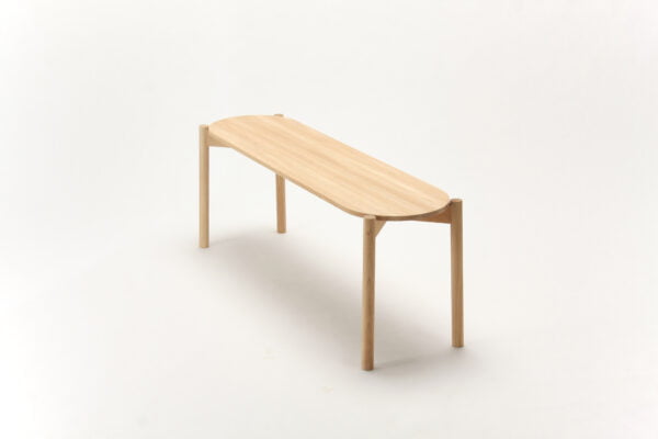 A picture of Sao A minimalist oak wooden bench with a light natural finish, featuring a long, narrow oval-shaped seat and four simple round legs. The bench is set against a plain, light-colored background.