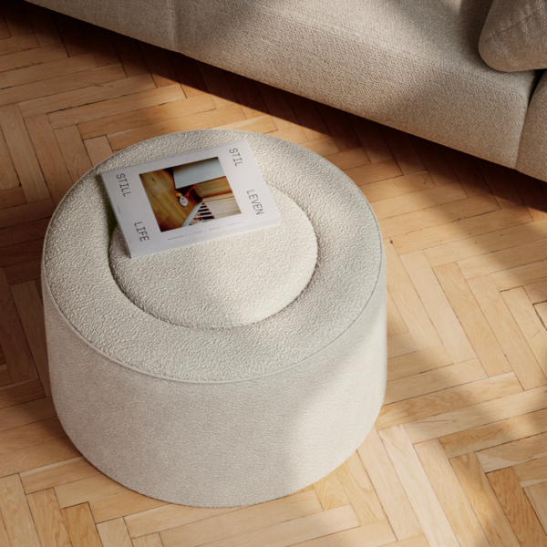 Top-down view of a circular, grey-colored Nel Pouf with textured fabric, placed beside a soft, neutral-toned sofa on wooden parquet flooring. A book titled 'Still Life' rests atop the Nel Pouf, contributing to a calm and minimalist interior design