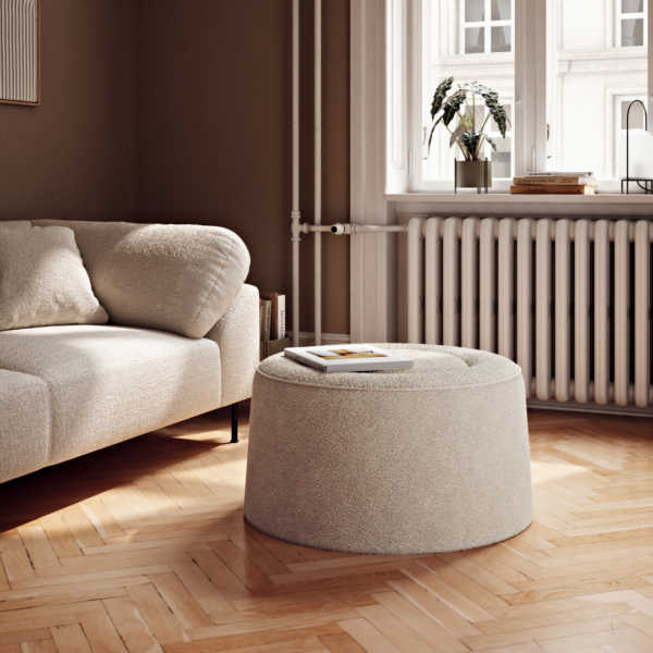 A cozy, minimalist living room featuring a neutral-colored, fabric-covered Nel Pouf placed next to a matching sofa. The Nel Pouf, in a circular shape, adds a touch of elegance to the wooden parquet flooring, illuminated by natural light streaming in from large arched windows. The overall decor is warm and serene, with a modern aesthetic