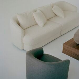 A cozy living room scene featuring the "Sophie" sofa in off-white, upholstered in boucle fabric. The curved design of the sofa is paired with matching cushions, adding a soft and inviting texture. A grey, rounded armchair is placed nearby, providing a subtle contrast to the light tones. The arrangement is completed with a wooden coffee table, enhancing the minimalist aesthetic of the room.
