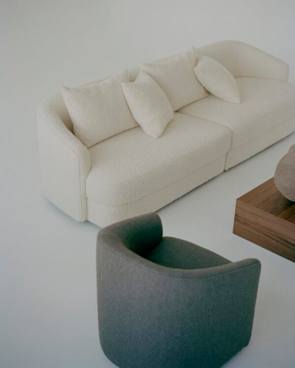 A cozy living room scene featuring the "Sophie" sofa in off-white, upholstered in boucle fabric. The curved design of the sofa is paired with matching cushions, adding a soft and inviting texture. A grey, rounded armchair is placed nearby, providing a subtle contrast to the light tones. The arrangement is completed with a wooden coffee table, enhancing the minimalist aesthetic of the room.