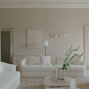 A serene and minimalist living room featuring the "Sophie" sofa, an off-white piece with elegant curved lines and boucle upholstery, crafted by Kersetna. The sofa is paired with matching cushions that highlight its inviting texture. The room is decorated in soft, neutral tones, with a large, plush rug underfoot and a sleek coffee table adorned with minimalist decor. A vase with tall green flowers adds a fresh touch, while subtle wall art and a sculptural light fixture enhance the calm, understated ambiance.