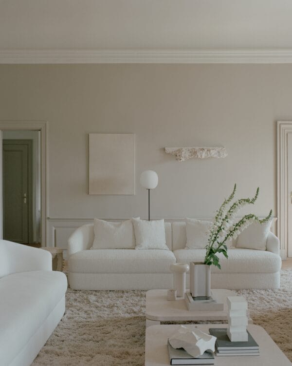 A serene and minimalist living room featuring the "Sophie" sofa, an off-white piece with elegant curved lines and boucle upholstery, crafted by Kersetna. The sofa is paired with matching cushions that highlight its inviting texture. The room is decorated in soft, neutral tones, with a large, plush rug underfoot and a sleek coffee table adorned with minimalist decor. A vase with tall green flowers adds a fresh touch, while subtle wall art and a sculptural light fixture enhance the calm, understated ambiance.